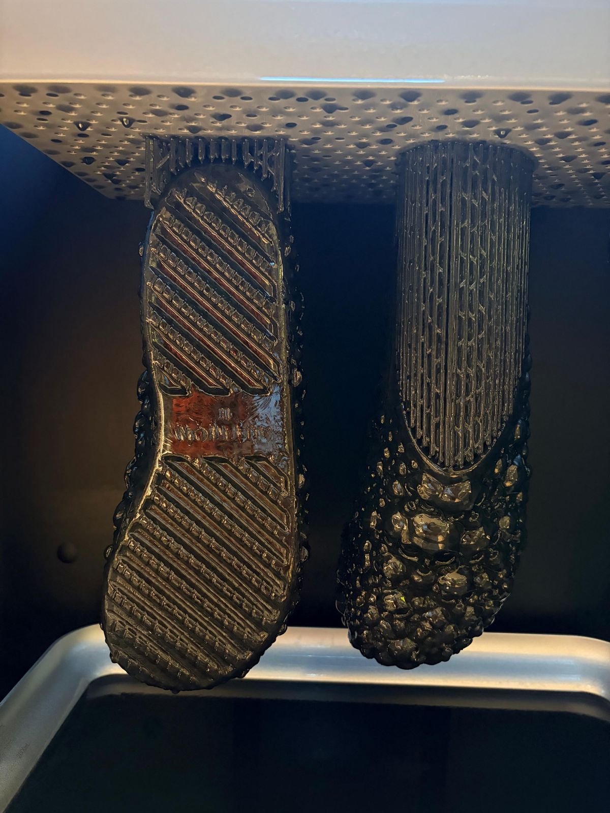 Two black resin-printed soles hanging from a printer's build platform.
