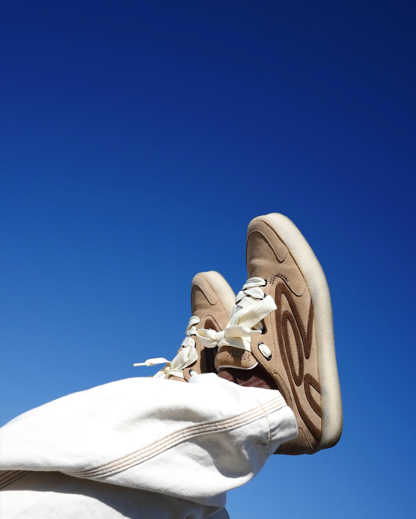 A pair of tan suede crepe sneakers with brown swirl overlays, worn against a blue sky.